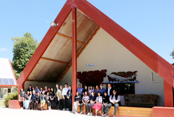 A large group of asian employee network staff sit outside the entrance of the marae